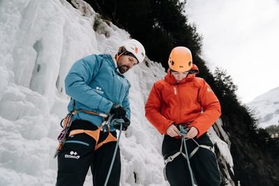 Two people with helmets and climbing harnesses stand in front of an icy rock wall in the mountains.