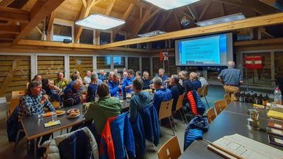 A group of people is sitting in a wooden room at tables while a presentation is being shown.