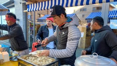 Personen servieren Essen an einem Marktstand mit blau-weißen Markisen im Hintergrund.
