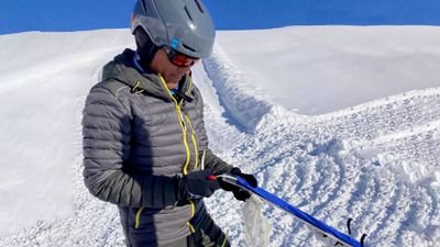 Person mit Helm und Skibrille hält Skistöcke im schneebedeckten Gebirge.