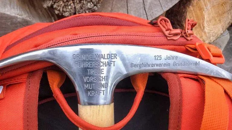 An ice axe with engraving is lying on a red backpack, with a woodpile in the background.