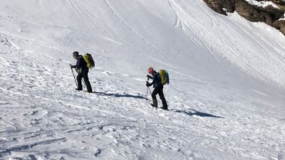 Zwei Personen mit Rucksäcken und Stöcken wandern im Schnee in einer bergigen Landschaft.