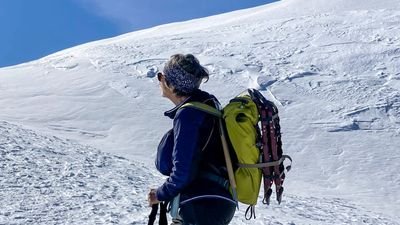 Person mit Rucksack und Wanderstöcken auf schneebedecktem Berg, trägt Stirnband und Sonnenbrille.