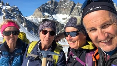 Four people with sunglasses and hats in front of snow-covered mountains, one of them holding an ice pick.