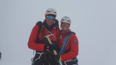Two people in climbing gear with helmets and ropes in a snow-covered mountain landscape.