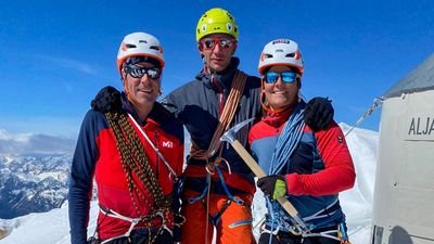 Three people with helmets and climbing equipment are standing on a snow-covered mountain peak.