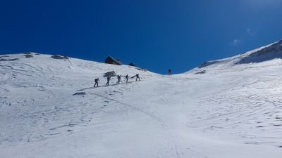 People with skis and poles are walking up a snowy mountain; clear blue sky.