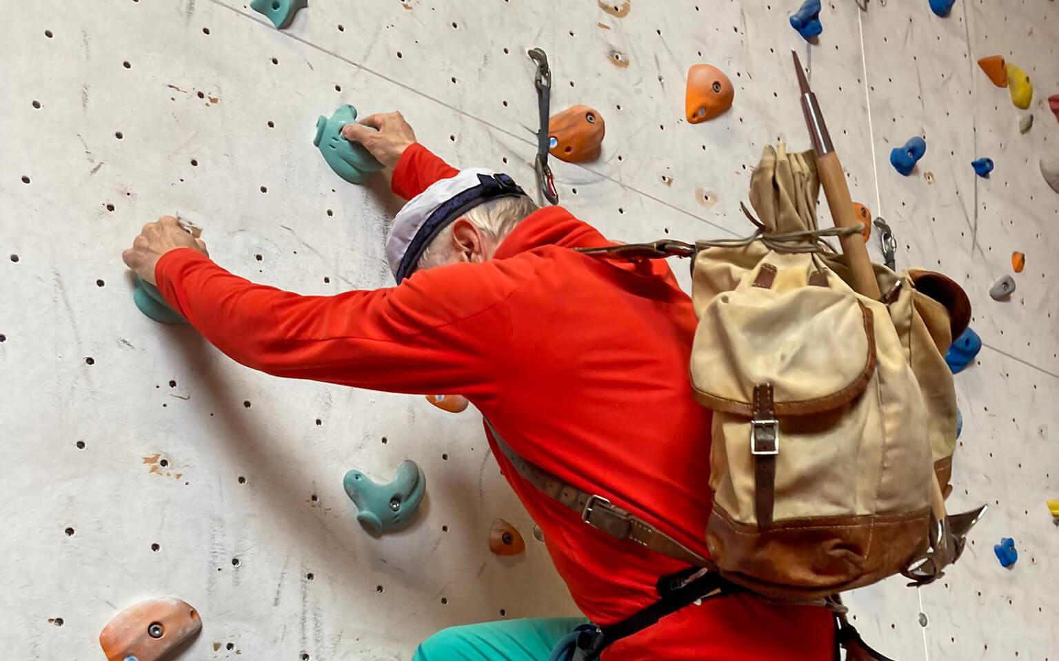 Person on climbing wall with helmet, harness and backpack in an indoor climbing gym.