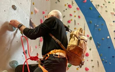 Person beim Klettern in einer Halle mit Klettergurt und Seil, bunte Griffe an der Wand.
