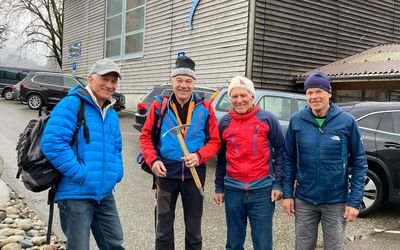 Vier Personen in Outdoor-Kleidung und Mützen stehen vor einem Gebäude mit Autos im Hintergrund.