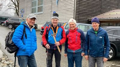 Four people in outdoor clothing and hats are standing in front of a building with cars in the background.