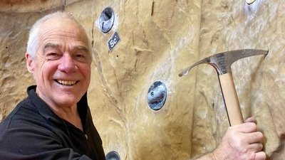 A person is holding an ice pick in front of a climbing wall in an indoor climbing gym.