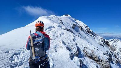 A person with a helmet and backpack on a snow-covered mountain ridge under a blue sky.