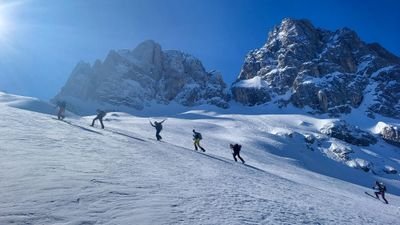 People with skis and backpacks are climbing a snowy mountain, with rocks in the background.