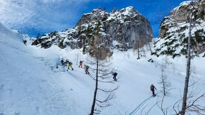 People with skis are skiing down a snow-covered slope, surrounded by mountains and trees.