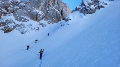 People with skis and backpacks are climbing a snowy mountain surrounded by rocks.