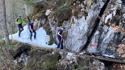 Three people with skis and backpacks are hiking on a narrow path along a rocky slope.