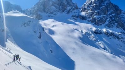 People with skis and helmets on a snowy mountainside, surrounded by high, snow-covered peaks.