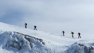 Five people with skis and backpacks are climbing up a snow-covered mountain.