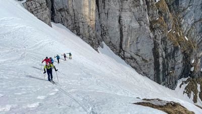 People with skis and backpacks are climbing up a snow-covered mountainside.