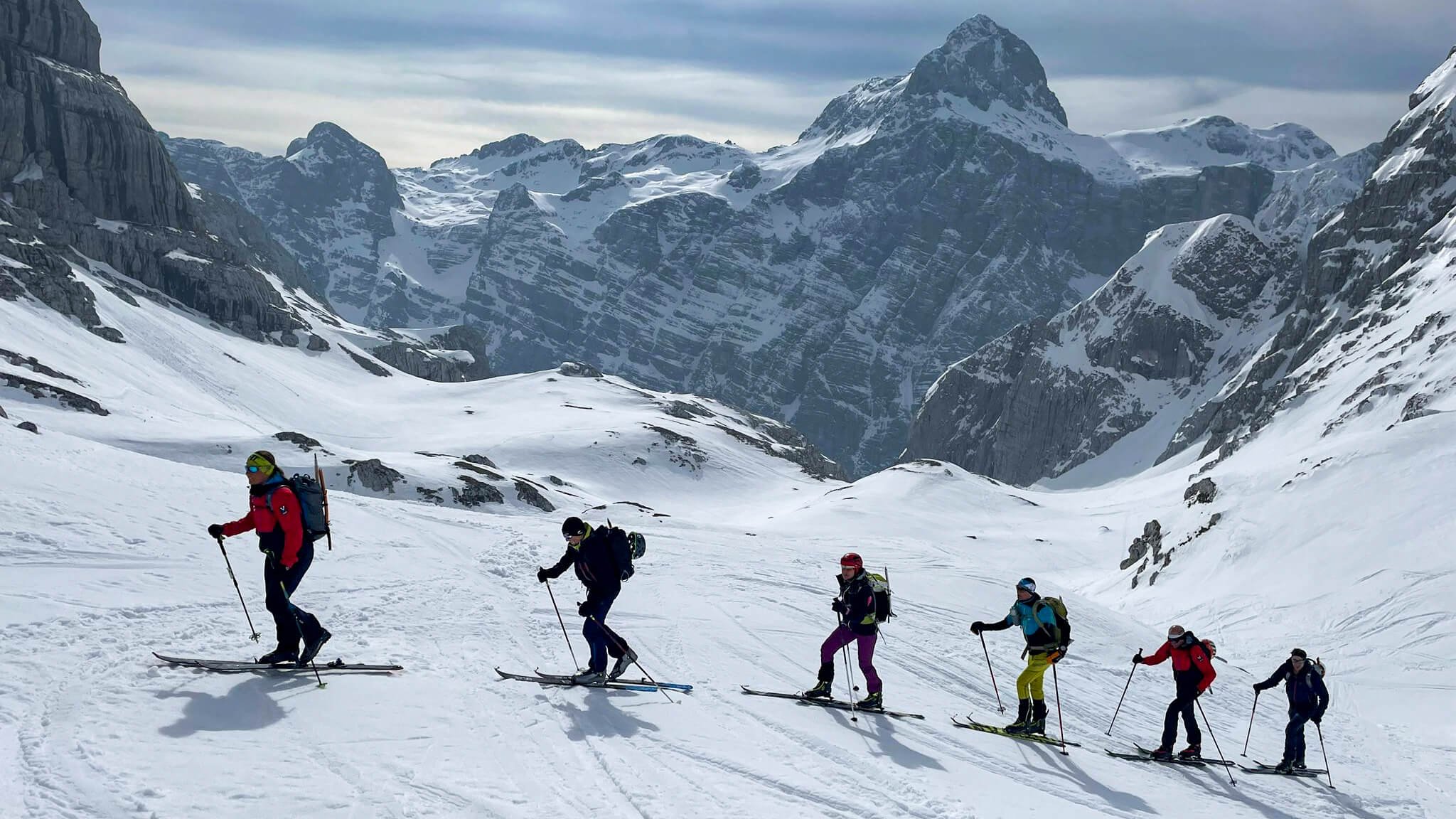 People with skis and backpacks are hiking in snowy mountains.