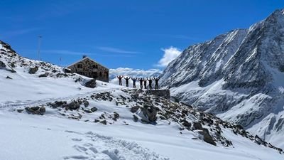 Personen stehen auf einer verschneiten Berglandschaft vor einer Hütte, umgeben von hohen Gipfeln.