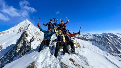 Gruppe von Personen mit Eispickeln auf schneebedecktem Berggipfel, umgeben von Alpenlandschaft.