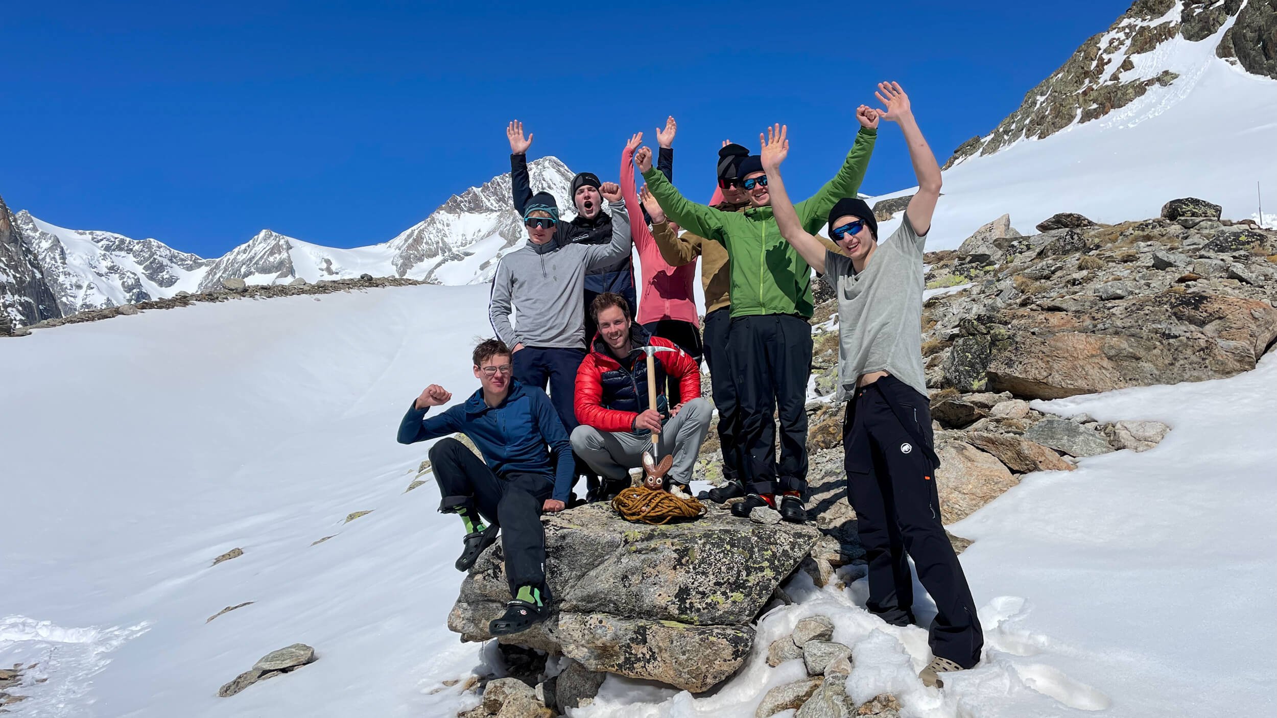 Group of people on a snow-covered mountain, some wearing sunglasses, raising their arms.