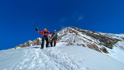 Personen mit Helmen und Kletterausrüstung wandern auf schneebedecktem Bergpfad unter blauem Himmel.