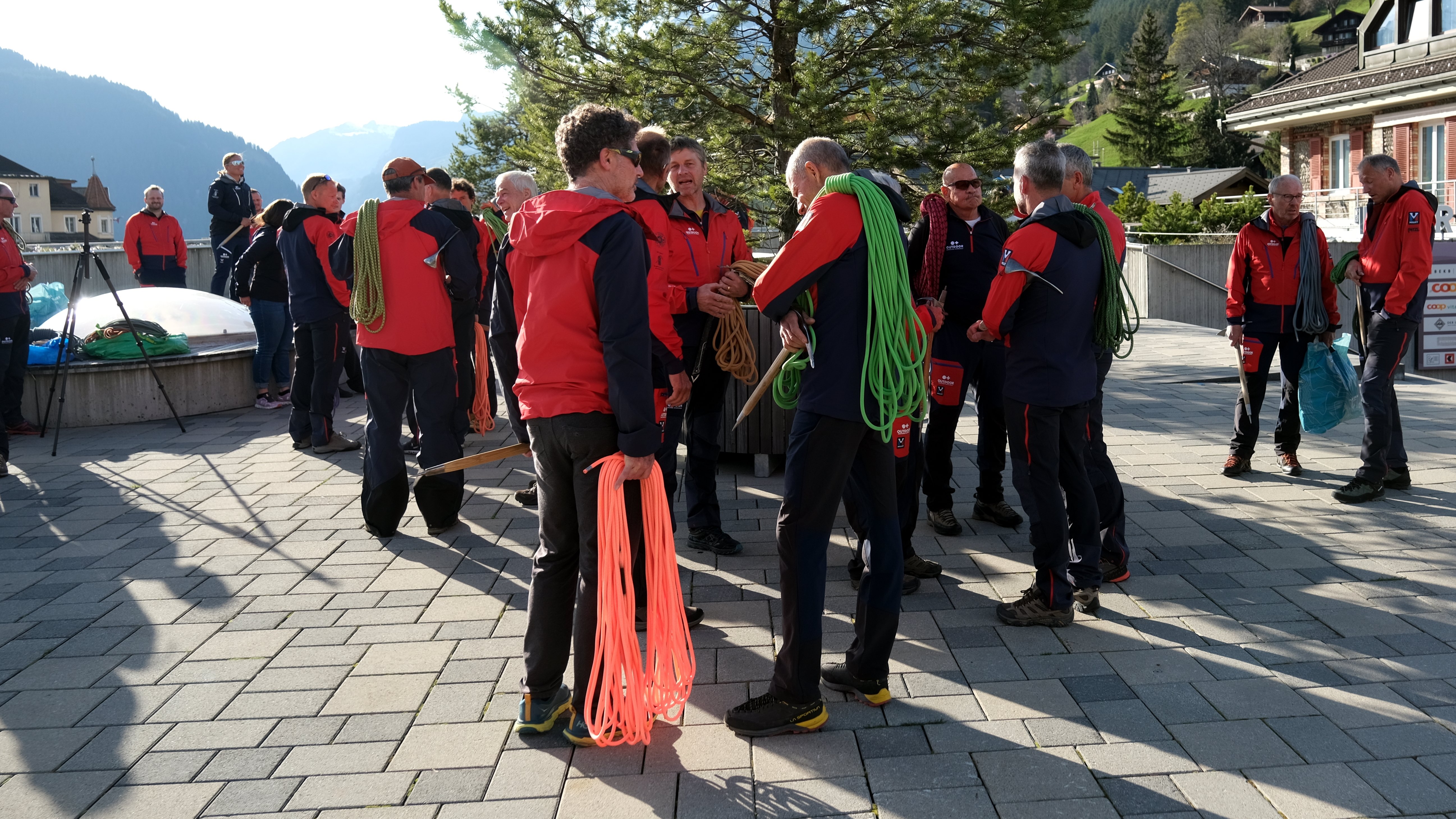Group of people in red jackets with climbing ropes on a paved square in the mountains.