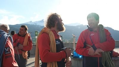 People in red outdoor clothing with ropes on a terrace, mountains in the background.