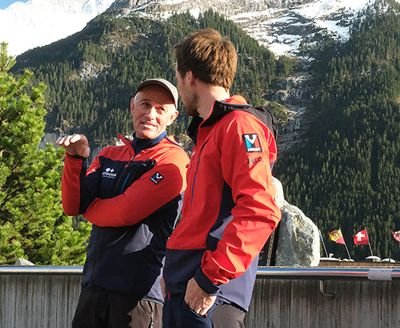 Two people in red jackets are standing in front of a mountain backdrop and talking to each other.