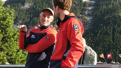 Two people in red jackets are standing in front of a mountain backdrop and talking to each other.
