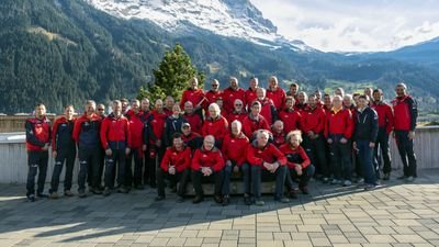 A group of people in red jackets is standing in front of a mountain backdrop on a terrace.