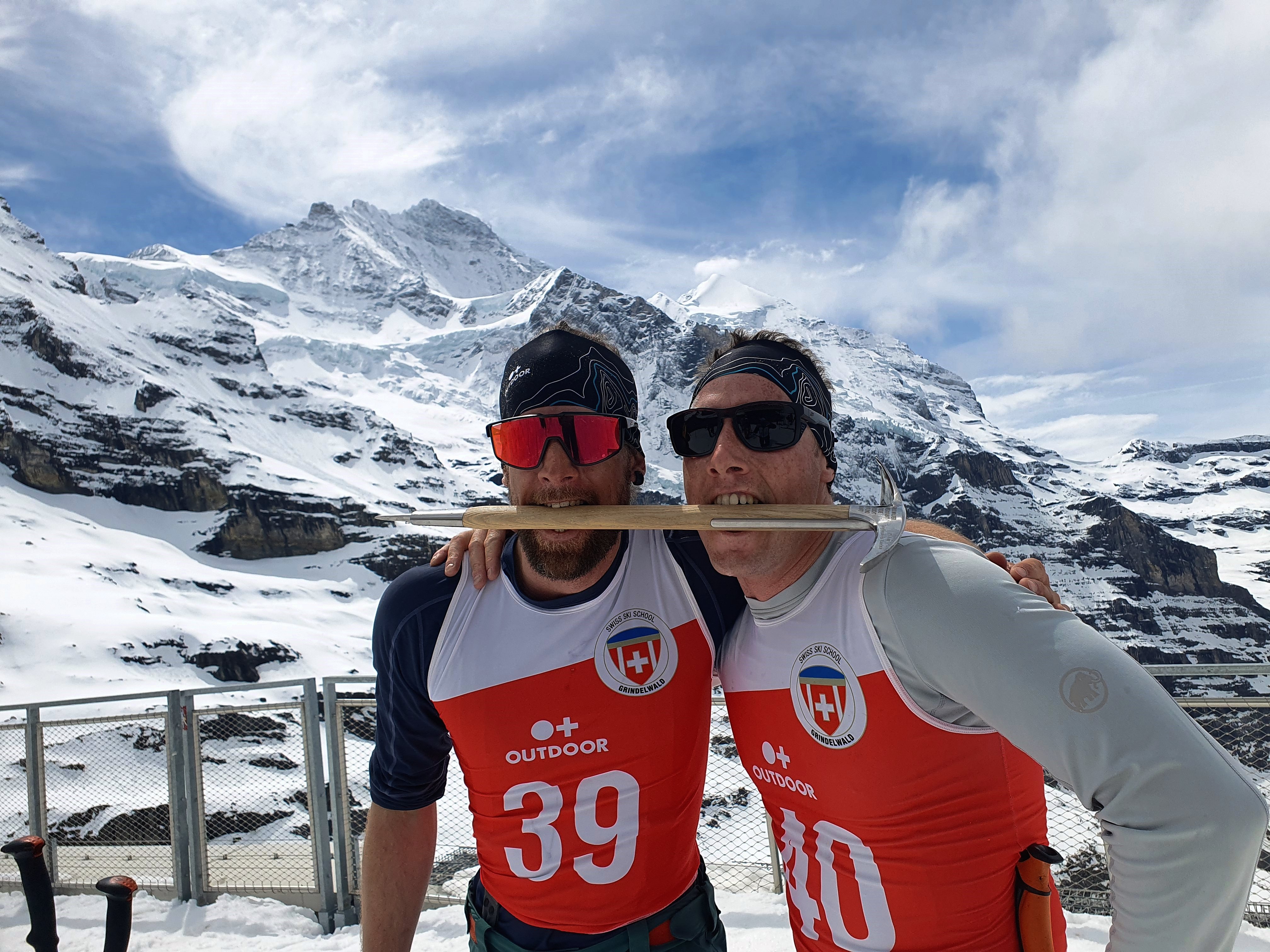 Two people in sports clothing with race numbers are standing in front of snow-covered mountains on a platform.