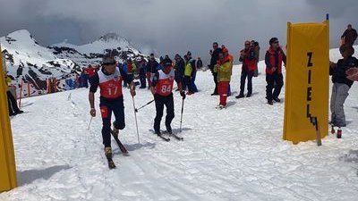 Two skiers with race numbers and poles pass a finish banner in the snowy mountains.