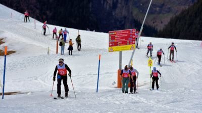 Personen mit Skiern und Helmen auf verschneiter Piste, rote Startnummern, Hinweisschilder im Hintergrund.