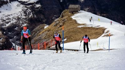 Drei Personen mit Startnummern und Skiern steigen einen schneebedeckten Hang in den Bergen hinauf.