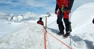 People with climbing equipment and ropes on a snow-covered mountain peak, surrounded by the Alps.