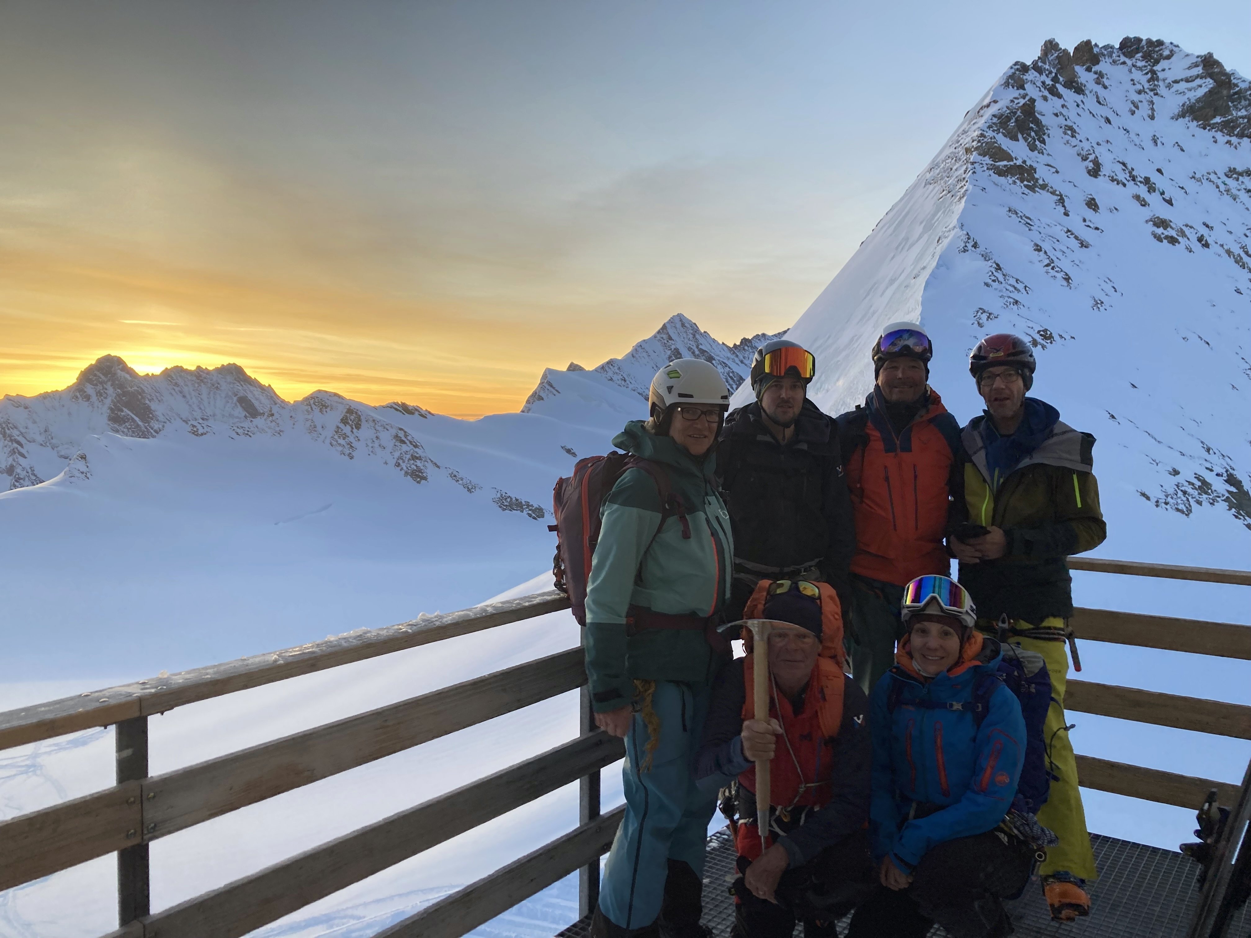 Group with helmets and ski equipment on wooden platform in front of snow-covered mountains at sunset.