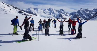 Gruppe von Skifahrern mit Helmen und Stöcken auf schneebedecktem Berg vor Alpenkulisse.