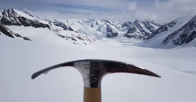 Eispickel vor schneebedeckter Berglandschaft mit Gletschern und bewölktem Himmel im Hintergrund.