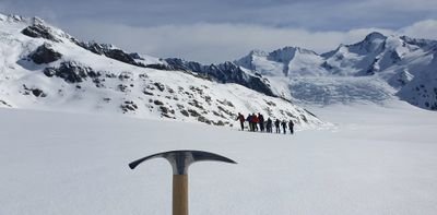 Gruppe von Menschen mit Bergsteigerausrüstung auf verschneitem Gletscher, Eispickel im Vordergrund.