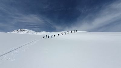 Group of people with ski equipment hiking in a snowy landscape, mountains in the background.