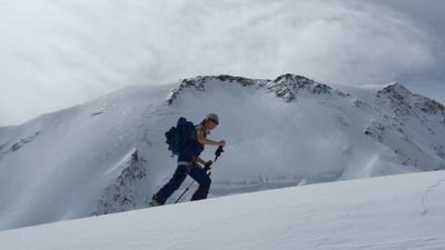 Person mit Helm und Rucksack steigt mit Skistöcken einen schneebedeckten Berg hinauf.