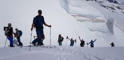 Group of people with sticks hiking in the snow in front of a glacier wall.