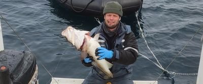 Person in outdoor clothing holding a large fish on a boat, with water and an inflatable boat in the background.