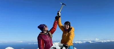 Two people in winter clothing on a snowy mountain peak, holding ice axes in the air.