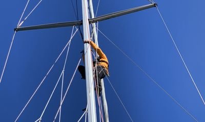Person in yellow jacket climbing on a sailboat mast, secured with a climbing harness, against a blue sky.