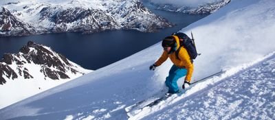 Person skiing down a snow-covered slope, with backpack and helmet, mountain landscape in the background.